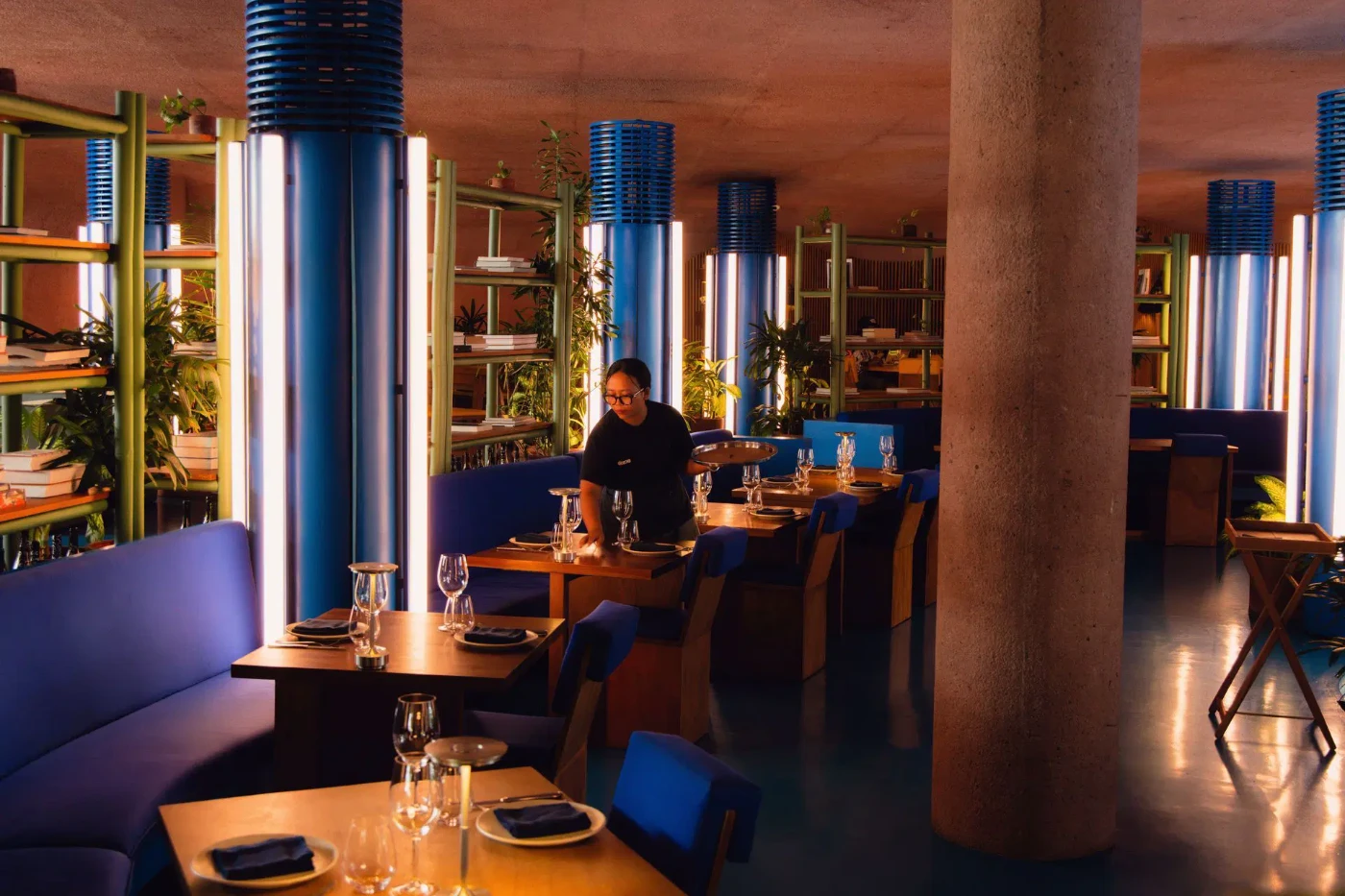 A server laying up tables in a concept restaurant lit by blue and green tube lighting.