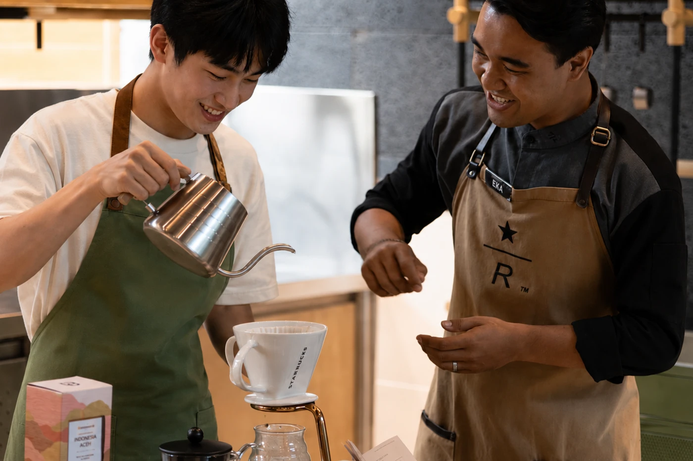 Two baristas in aprons brewing a pour-over together behind a counter.