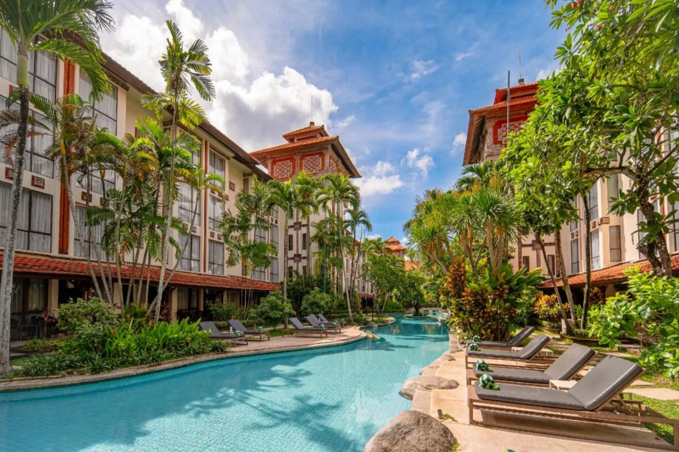 A hotel pool running between two low wings, with palms, sun loungers and pitched tile roofs.